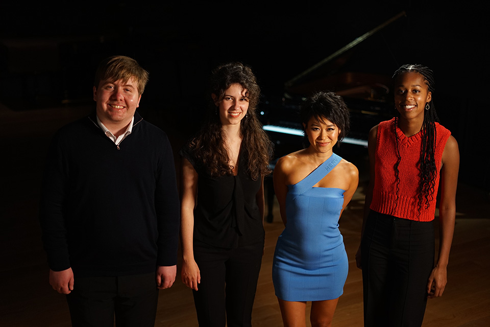 Four people, one male, three female, standing in front of a piano in a dark room